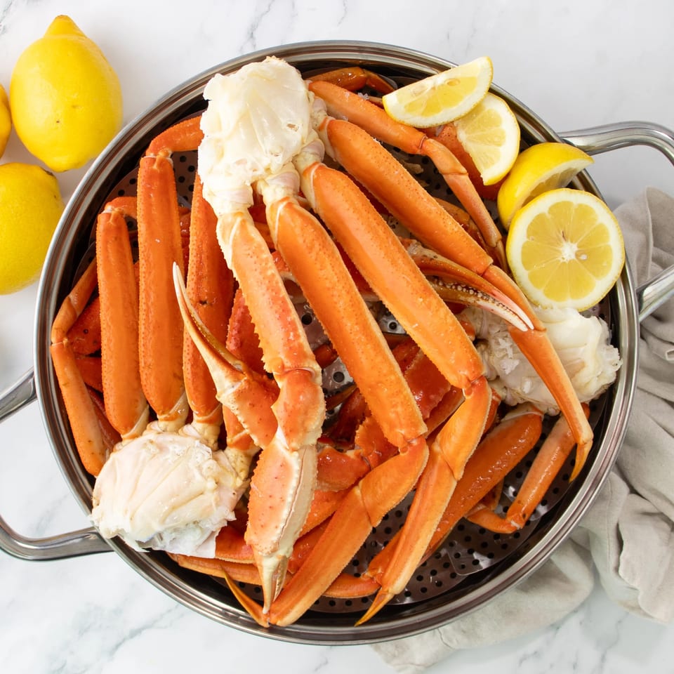 Steamed crab legs and claws with lemon wedges in a metal steamer basket on a marble surface.