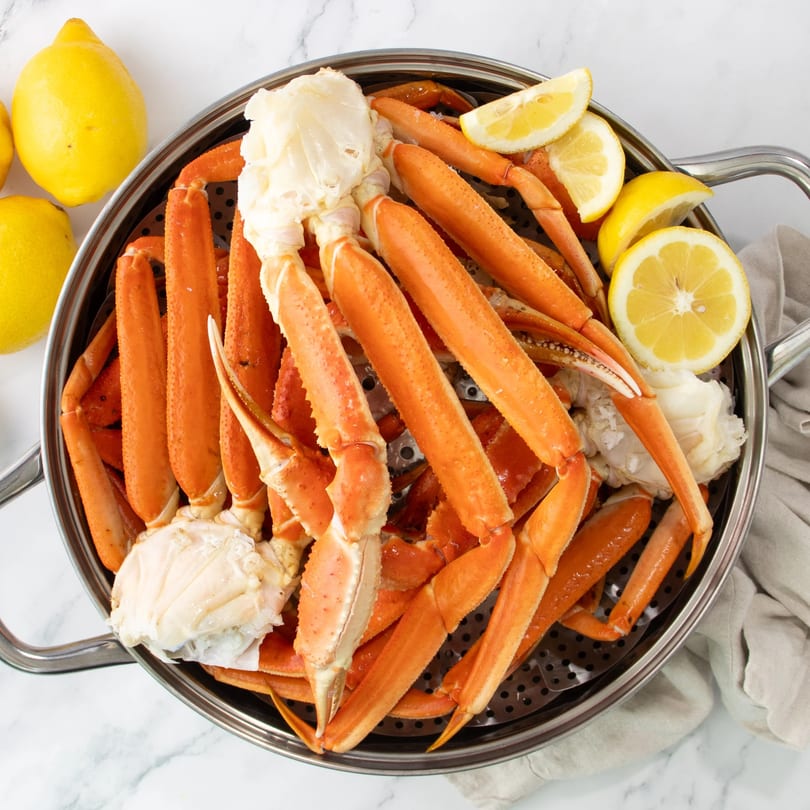 Steamed crab legs and claws with lemon wedges in a metal steamer basket on a marble surface.