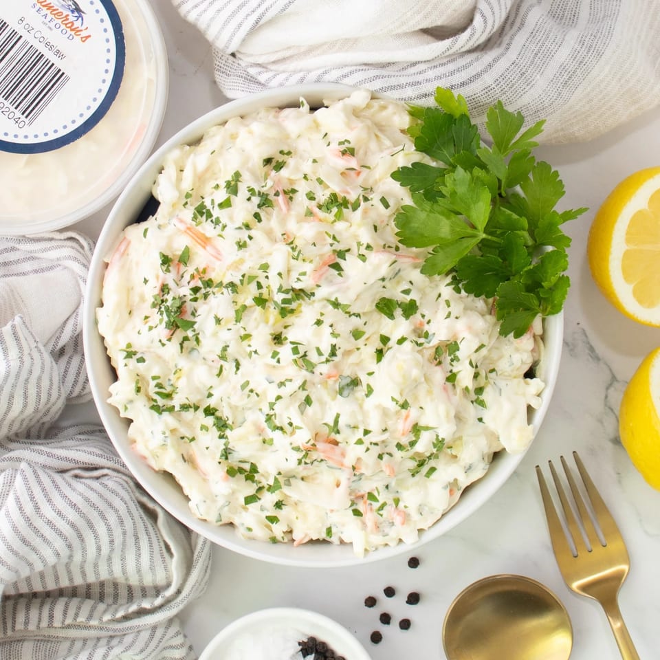 Creamy coleslaw in a white bowl, garnished with parsley, next to lemons and a container of Cameron's Seafood coleslaw.