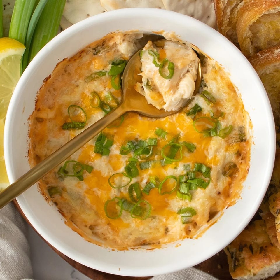 Golden spoon scooping creamy crab dip with melted cheese and green onions, served with bread and lemon.