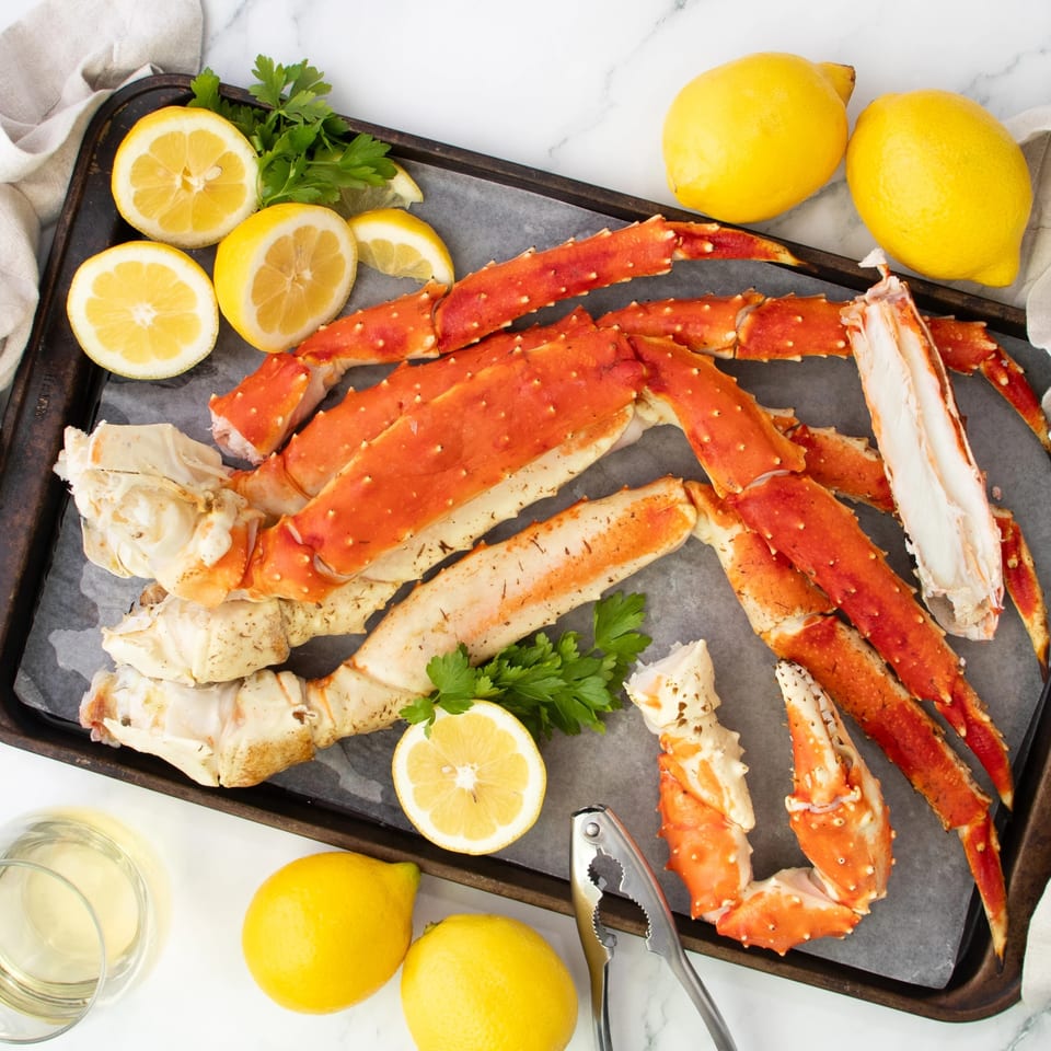 Cooked king crab legs, lemons, parsley, and a nutcracker on a dark baking sheet.
